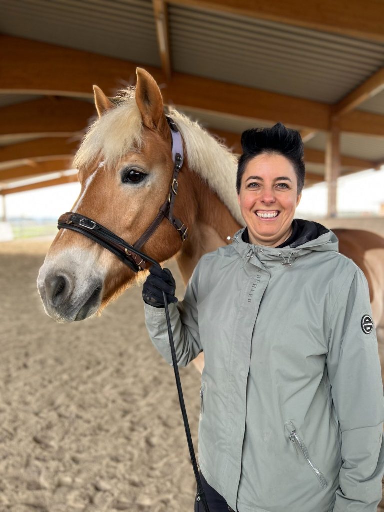 Reiter longiert ein Pferd in einer sandigen Reitarena mit Holzdach.